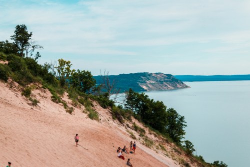 A sandy beach with people enjoying the sun, overlooking a calm lake and distant hills under a blue sky.