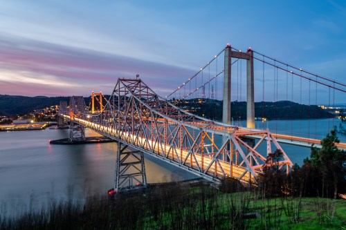 A bridge spans a river at dusk, illuminated by lights, with a colorful sky and hills in the background.