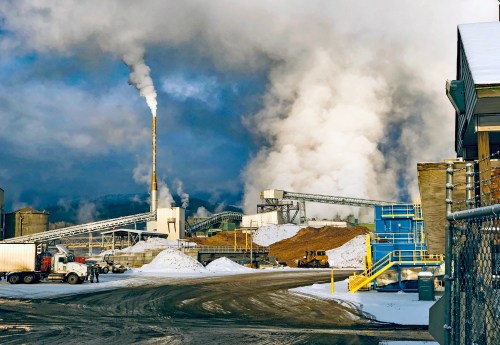 Industrial site with smokestacks, machinery, and snow-covered ground under a cloudy sky.