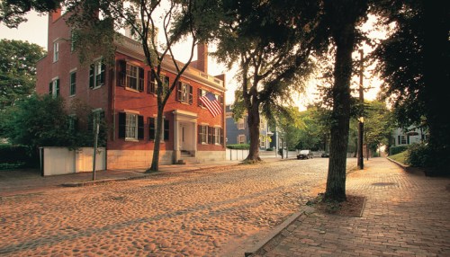 A historic brick building with an American flag, surrounded by trees and a cobblestone street at sunset.
