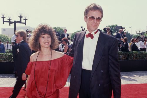 A couple poses on the red carpet at an outdoor event, surrounded by cameras and onlookers, in stylish attire.