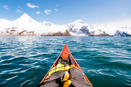 A kayak glides over turquoise water, with snow-capped mountains rising in the background under a clear blue sky.