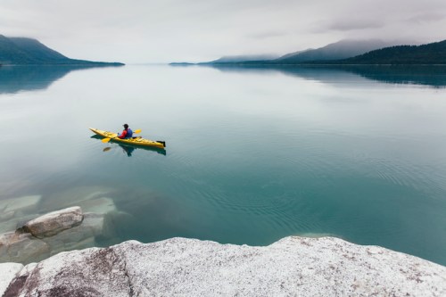 A person in a yellow kayak paddles on a calm lake surrounded by mountains under a cloudy sky.