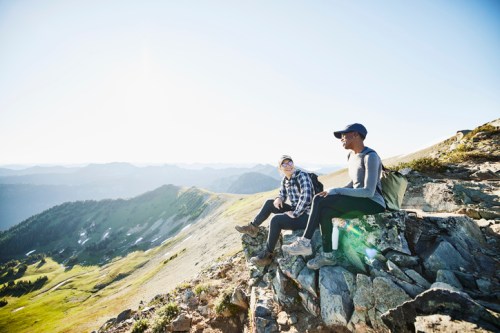 Two hikers sit on a rocky outcrop, enjoying the scenic mountain view under a clear blue sky.