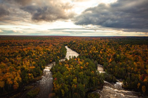 Aerial view of a winding river surrounded by vibrant autumn foliage and dramatic clouds in the sky.
