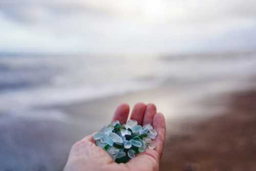 A hand holding a collection of colorful sea glass against a blurred beach and ocean background.