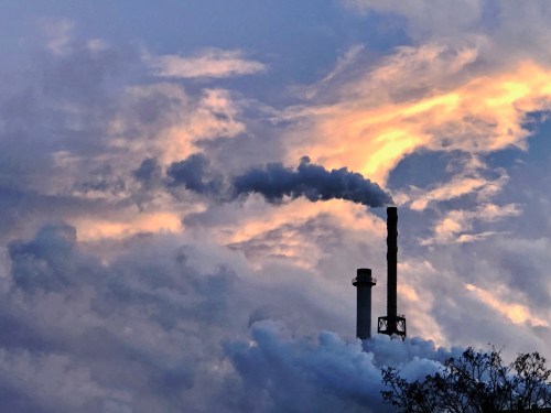 A factory smokestack emits smoke against a backdrop of dramatic clouds and a colorful sunset.