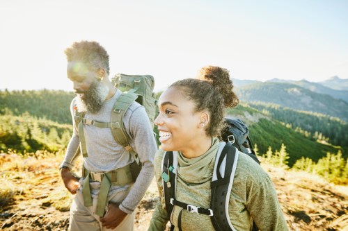 Two hikers smile while trekking through a scenic mountain landscape, surrounded by trees and sunlight.