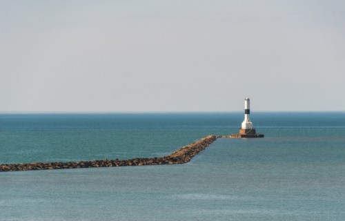 A lighthouse stands at the end of a rocky jetty, surrounded by calm blue waters under a clear sky.