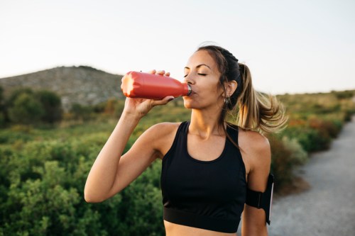 A woman in a black sports top drinks from a red water bottle while jogging outdoors.