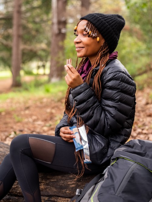 A woman in a black puffer jacket and beanie sits on a log, enjoying a snack in a forested area.