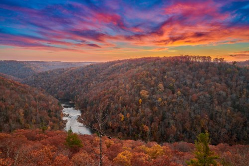 A vibrant sunset over rolling hills covered in autumn foliage, with a winding river below.
