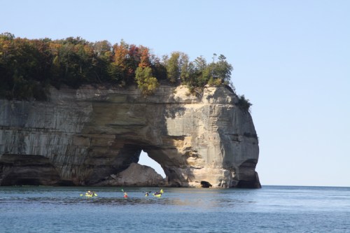 A scenic view of a large rock formation with an arch, surrounded by calm water and autumn-colored trees.