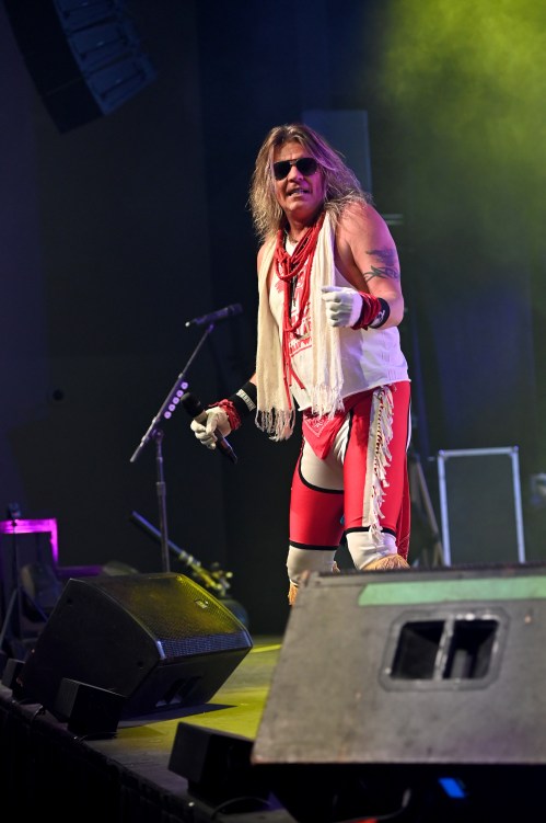 A performer in a red outfit and sunglasses sings on stage, with colorful lights and musical equipment in the background.