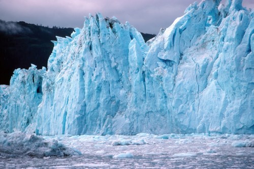 A close-up of a towering blue glacier with jagged ice formations against a cloudy sky.