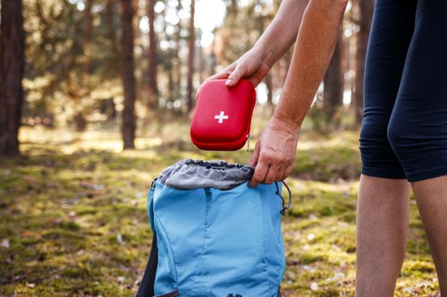A person places a red first aid kit into a blue backpack in a forested area.