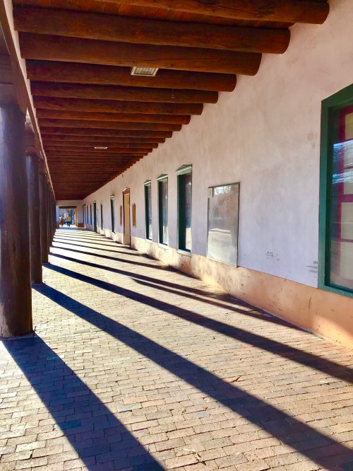 A sunlit corridor with wooden beams and long shadows cast on a brick path beside white walls and green windows.