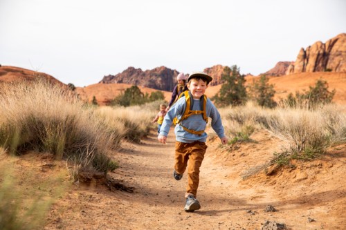A young boy runs joyfully on a dirt trail, with two children and rocky terrain in the background.