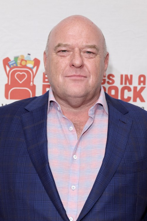 A bald man in a blue blazer and pink shirt smiles at the camera, standing in front of a promotional backdrop.