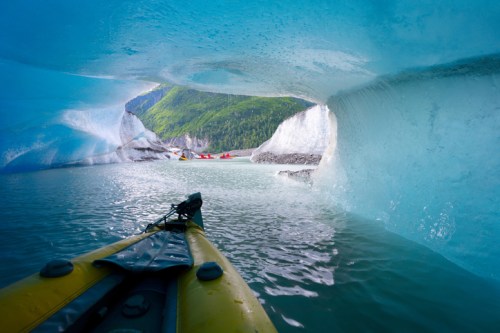 A kayak view through an ice arch, revealing a serene glacial landscape and other kayakers in the distance.