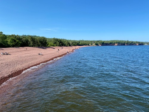A serene beach with a pebbly shore, surrounded by green trees and calm blue water under a clear blue sky.
