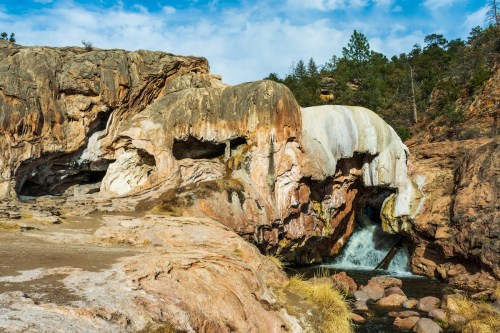A rocky landscape featuring a waterfall cascading through a natural stone formation under a blue sky.