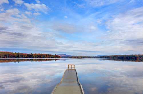 A serene lake view with a wooden dock extending into calm waters, surrounded by colorful autumn foliage and a blue sky.