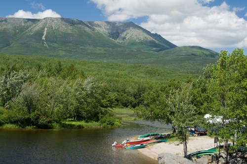 A serene landscape featuring a riverbank with colorful boats, lush greenery, and a mountain under a blue sky with clouds.
