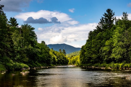 A serene river flows between lush green trees, with mountains and a partly cloudy sky in the background.