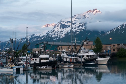 A serene harbor with fishing boats, surrounded by snow-capped mountains and a cloudy sky.