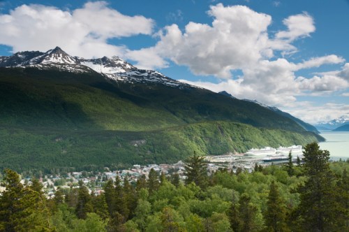 A scenic view of a coastal town surrounded by lush green forests and snow-capped mountains under a blue sky with clouds.