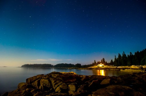 A serene night scene with stars above a calm sea, rocky shore, and a distant house illuminated by warm light.