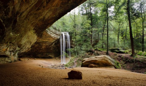 A serene forest scene featuring a waterfall cascading from a rocky overhang into a sandy area surrounded by trees.
