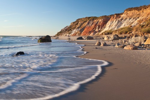 A serene beach scene with gentle waves lapping at the shore, rocky cliffs in the background under a clear blue sky.