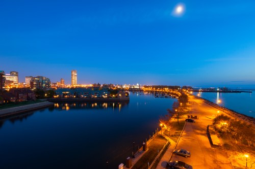 A serene waterfront view at dusk, featuring city lights reflecting on calm water and a clear blue sky.