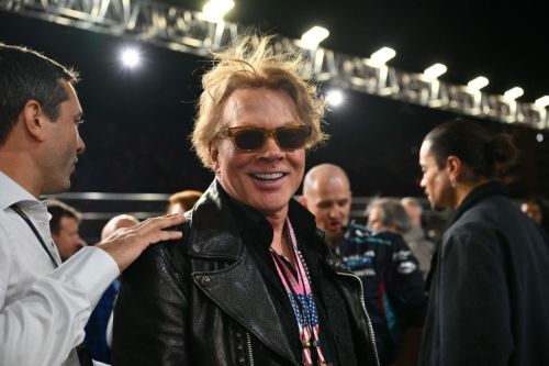 A smiling man with sunglasses and messy hair, wearing a leather jacket, stands among a group of people at an event.