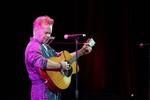 A musician in a blue jumpsuit plays guitar and harmonica on stage, illuminated by colorful lights.