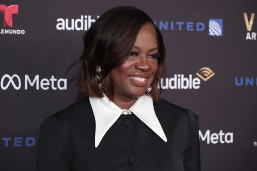 Viola Davis smiles at an event, wearing a black outfit with a white collar, against a branded backdrop.
