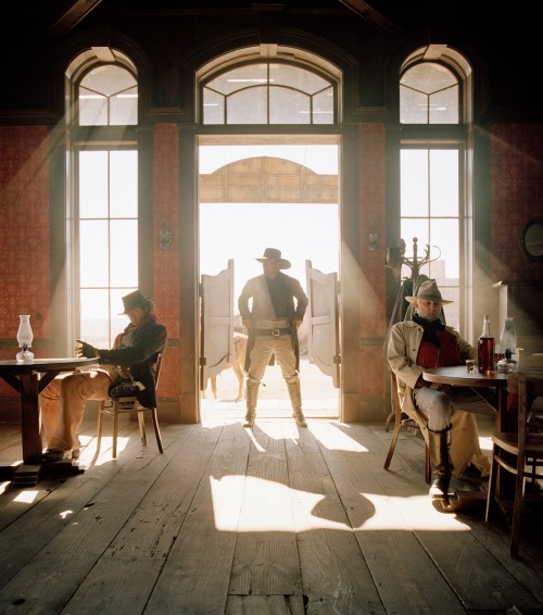 A cowboy stands in a doorway, with two seated figures in a sunlit, rustic interior of an old saloon.