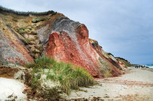 Colorful cliffs rise above a sandy beach, with people walking in the distance under a cloudy sky.