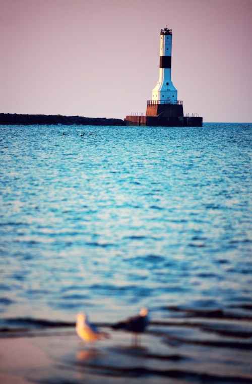 A lighthouse stands on a rocky pier, surrounded by calm blue water and a soft sunset glow in the background.