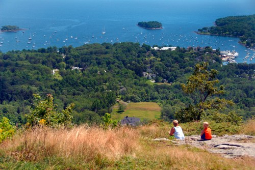 Two children sit on a rocky outcrop, overlooking a lush landscape and a bay filled with boats.