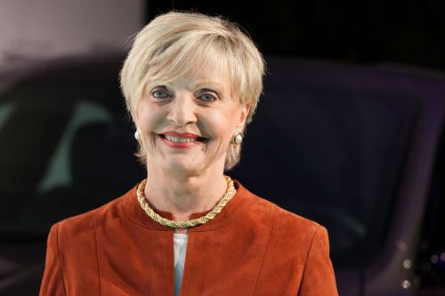 A smiling woman with short blonde hair, wearing a brown suede jacket and a gold necklace, stands in front of a dark background.