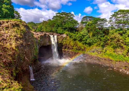A stunning waterfall cascades into a pool, surrounded by lush greenery and a rainbow in the mist under a blue sky.
