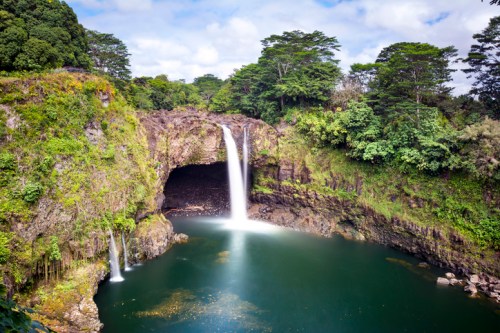 A serene waterfall cascades into a tranquil pool, surrounded by lush greenery and rocky cliffs under a blue sky.