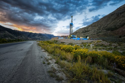 A drilling rig stands beside a road, surrounded by wildflowers and mountains under a cloudy sunset sky.