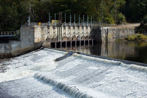 A dam with water cascading over its edge, surrounded by trees and a calm river.
