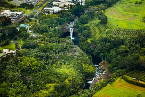 Aerial view of lush green landscape with a waterfall, river, and nearby buildings amidst dense vegetation.