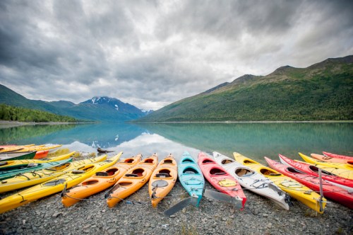 Colorful kayaks lined up on a rocky shore, reflecting in calm water with mountains and cloudy sky in the background.