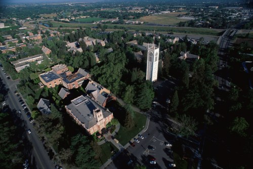 Aerial view of a campus with buildings, trees, and a tall clock tower surrounded by green fields and roads.